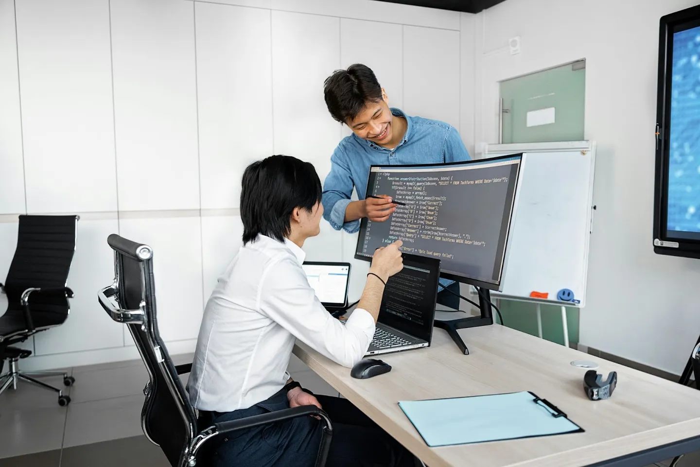 Test Driven Development vs Behavior Driven Development One young adult man sitting in front of large monitor with code and another young adult male standing, pointing at screen